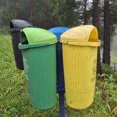Gekleurde afvalbakken voor gescheiden afval langs een wandelpad op de Azoren: groen voor glas, geel voor plastic, blauw en zwart op de achtergrond, omringd door bos