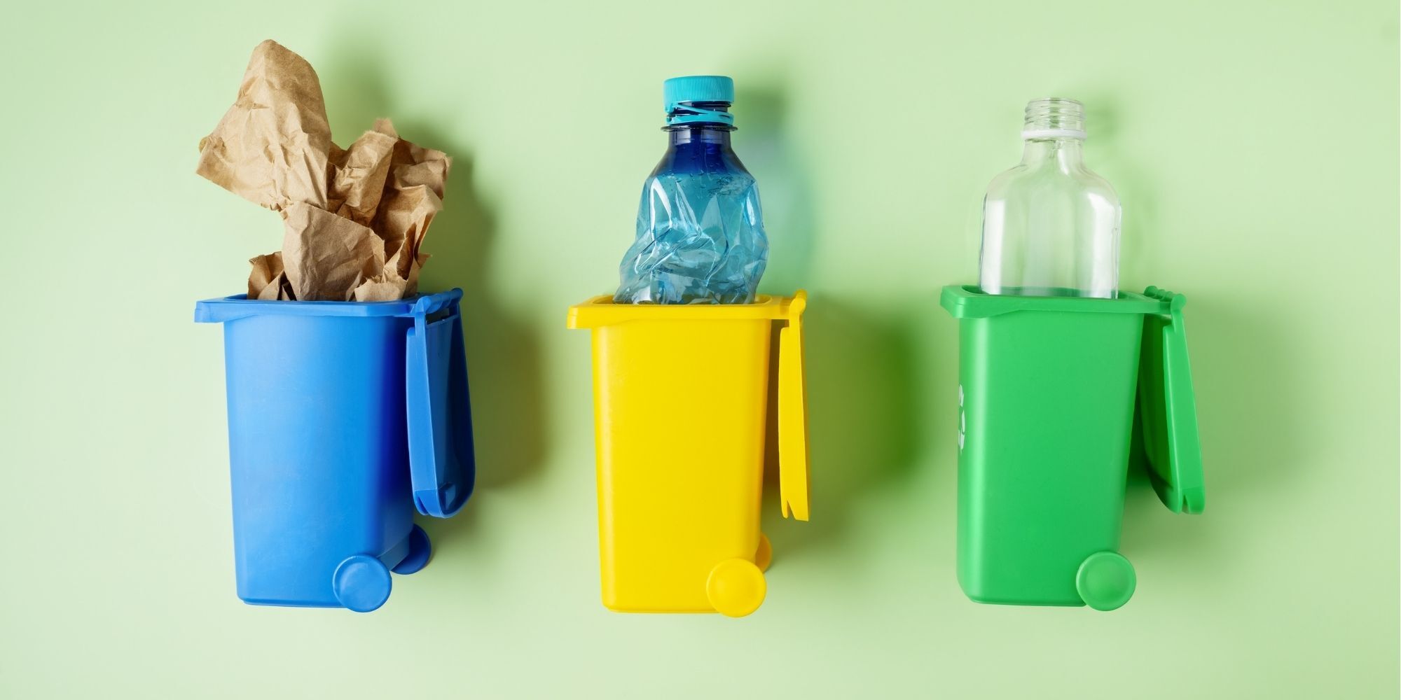 Drie kleine afvalbakken in blauw, geel en groen met daarin papier, een plastic fles en een glazen fles, ter illustratie van afvalscheiding in de juiste bak op de werkvloer.